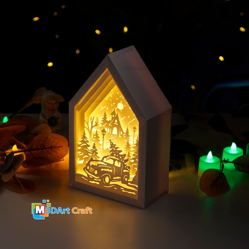 May include: A white wooden house-shaped shadow box with a cut-out design of a winter scene. The scene includes a car, a church, trees, and a full moon. The box is lit from the inside, creating a warm glow.