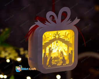 May include: White papercraft ornament shaped like a gift box with a red ribbon. The box has a cut-out design of the nativity scene with a star and palm trees. The ornament is illuminated from within.