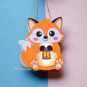 Op de afbeelding: Een hangende lantaarn in de vorm van een vos, gemaakt van oranje, wit en zwart papier. De vos heeft een wit gezicht, roze wangen en zwarte ogen, en houdt een kleine bruine lantaarn vast met een gele steruitsparing. Het midden geeft licht.