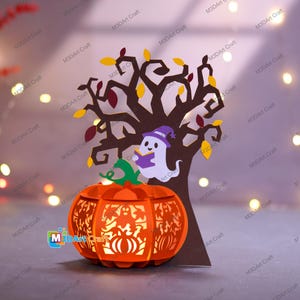 May include: A Halloween-themed papercraft featuring a carved orange pumpkin lantern with a tree and a ghost reading a book. The tree has brown branches with yellow and red leaves. The ghost wears a purple witch hat. The lantern has cut-out designs.