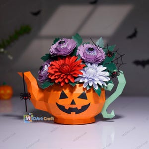 Może przedstawiać: Pomarańczowy czajniczek z papieru typu low-poly z czarną twarzą dyni, wypełniony papierowymi kwiatami w kolorach czerwonym, fioletowym i białym. Czajniczek posiada zielone liście, pajęczynę i małe czarne pająki. Widoczne jest logo M3DArt Craft.