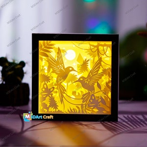 May include: A 3D paper cut-out lamp with a black frame. The design features two hummingbirds flying in a jungle scene with a moon and stars. The lamp is lit from behind, creating a warm glow.