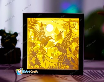 May include: A 3D paper cut-out lamp with a black frame. The design features two hummingbirds flying in a jungle scene with a moon and stars. The lamp is lit from behind, creating a warm glow.
