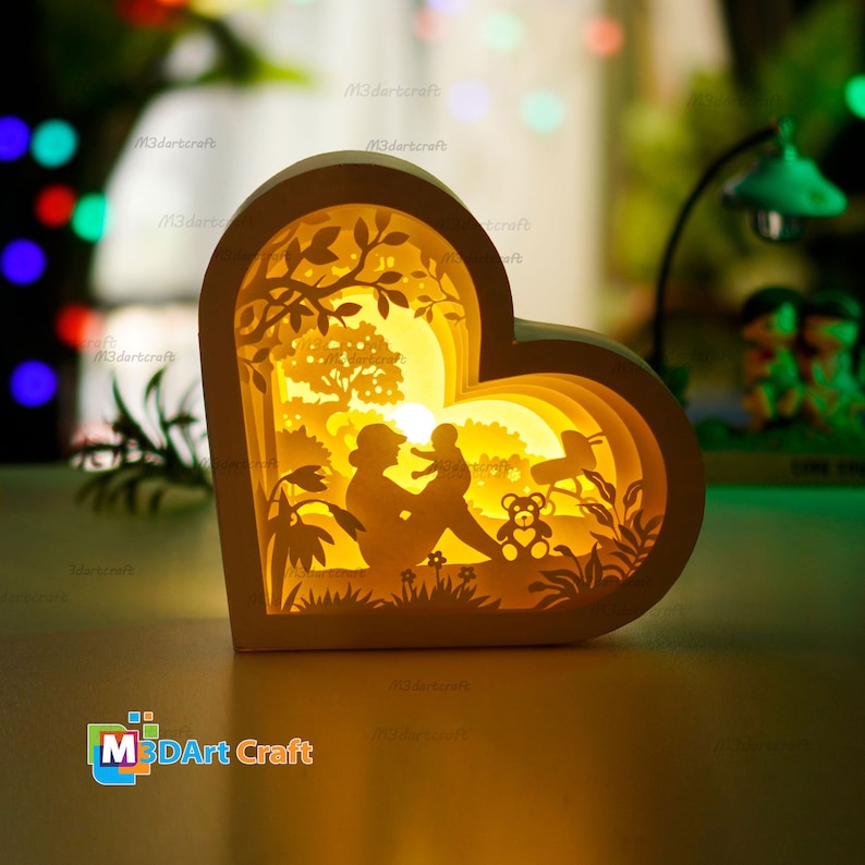 May include: A white wooden heart-shaped lamp with a 3D layered design. The design features a mother and child sitting in a park with a teddy bear. The lamp is illuminated with warm yellow light.