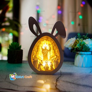 Può includere: Una lampada di legno bianca a forma di uovo di Pasqua con orecchie di coniglio. L'uovo è illuminato dall'interno e presenta un design di taglio dettagliato di Gesù Cristo e dei suoi discepoli. La lampada è appoggiata su un tavolo con uno sfondo sfocato.