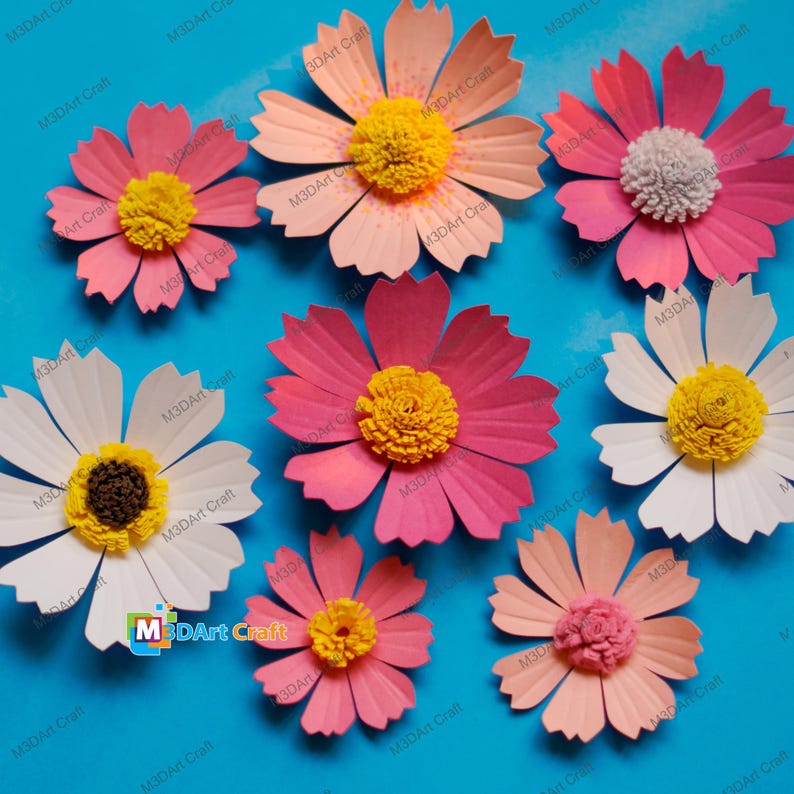 May include: A collection of handmade paper flowers in various shades of pink, peach, and white, with yellow or textured centers. The flowers are arranged on a bright blue background, showcasing the delicate petal details and craftsmanship. The text "M3DArt Craft" is visible.