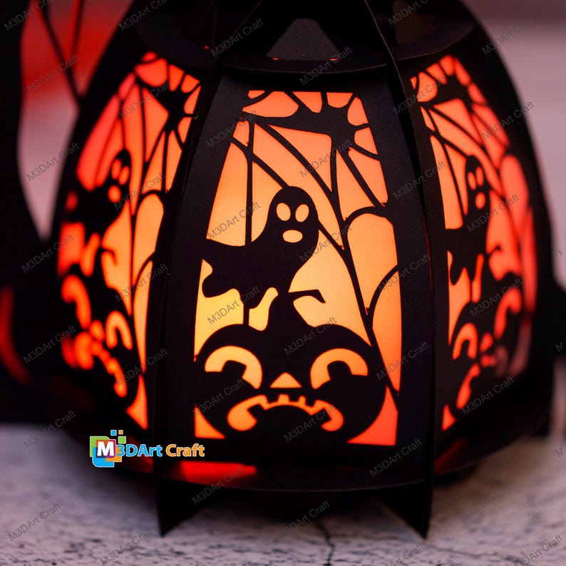 May include: A black paper lantern shaped like a pumpkin with a glow-in-the-dark design. The lantern features a ghost, spiderwebs, and a pumpkin face. The lantern is lit from inside, creating a warm orange glow.