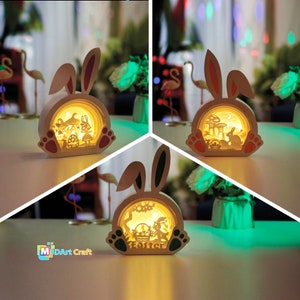 May include: Three white bunny shaped night lights with a 3D Easter scene inside. The lights are illuminated with a warm yellow glow. The bunny on the left has red ears, the bunny in the middle has green ears, and the bunny on the right has red ears. The bunny on the left has the text "M3DArt Craft" on the base. The bunny in the middle has the text "Easter" on the base.
