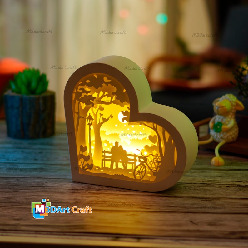 May include: A white wooden heart-shaped lamp with a cutout design of a couple sitting on a bench under a tree with a bicycle. The lamp is lit up with a warm yellow light.