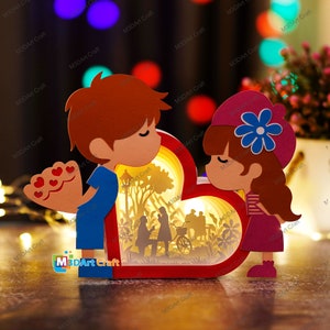 May include: A 3D papercraft model of a couple in love. The couple is standing in front of a heart-shaped window with a scene of a park inside. The model is made of red, pink, blue, and white paper.