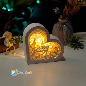 May include: A white heart-shaped wooden lamp with a 3D layered design of a sea turtle swimming in the ocean. The lamp is lit up from the inside, creating a warm glow.