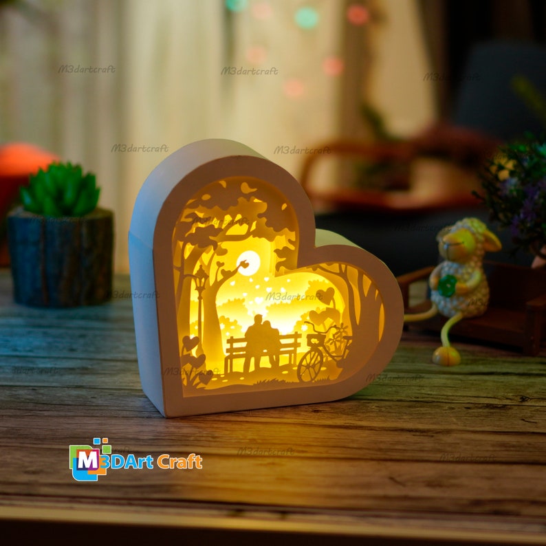 May include: A white heart-shaped wooden lamp with a cutout design of a couple sitting on a bench in a park. The lamp is illuminated from the inside, creating a warm glow. The text "M3DArt Craft" is visible at the bottom of the image.