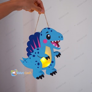 May include: A blue dinosaur-shaped hanging light with dark blue spots, a pink cheek, and a yellow illuminated belly. The figure features a dark blue dorsal fin with pink stripes, a white-toothed open mouth, and black claws, suspended by a thin twine.