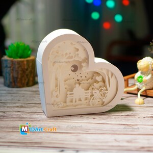 May include: White heart-shaped papercraft lamp with a 3D design of a couple sitting on a bench in a park with a bicycle and a moon in the background. The lamp is lit up and the design is visible through the cutouts.