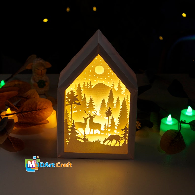 May include: A white wooden house-shaped nightlight with a cut-out design of a forest scene with deer, trees, and a moon. The light is illuminated from within, casting a warm glow.