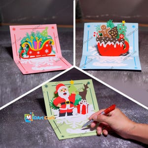 May include: Three pop-up Christmas cards. A pink card shows a sleigh with gifts. A blue card features a mug with gingerbread and pine cones. A green card displays Santa Claus with a Christmas tree and a gift. The cards have snowflake and star designs.