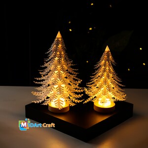 Peut inclure: Deux sapins de No&euml;l en papier blanc avec des lumi&egrave;res &agrave; l'int&eacute;rieur, pos&eacute;s sur une base noire. Les arbres sont &eacute;clair&eacute;s de l'int&eacute;rieur, cr&eacute;ant une lueur chaleureuse. La base porte l'inscription "M3DArt Craft".