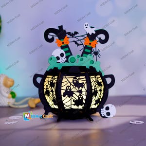 Puede incluir: Un caldero negro con la parte superior verde y un interior iluminado. El caldero está decorado con una telaraña, fantasmas y piernas de bruja. Hay dos calaveras en el caldero.
