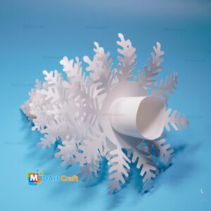 Peut inclure: Ornement en papier blanc en forme de flocon de neige avec un tube en papier blanc au centre. L'ornement est sur un fond bleu clair. Le texte "M3DArt Craft" est visible dans le coin inf&eacute;rieur gauche de l'image.
