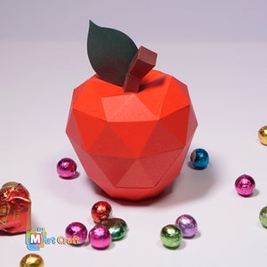 K&ouml;nnte beinhalten: Ein roter Papier-Apfel mit einem gr&uuml;nen Blatt oben. Der Apfel besteht aus gefaltetem Papier und hat ein geometrisches Design. Um den Apfel herum liegen kleine, bunte, runde Bonbons.