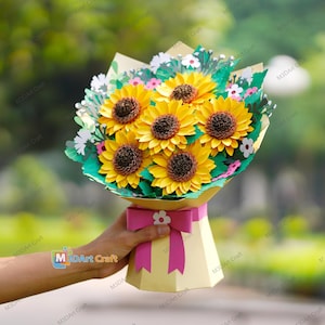 Può includere: Un bouquet di carta con sei girasoli gialli con centri marroni, circondati da foglie e fiori di carta verde e bianca. Il bouquet è avvolto in carta gialla e legato con un fiocco rosa.