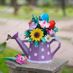 Könnte beinhalten: Eine lila Papier-Gießkanne mit weißen Punkten, gefüllt mit bunten Papierblumen. Eine gelbe Sonnenblume ist prominent, mit roten, rosa und blauen Blüten. Ein Papier-Schmetterling ruht auf dem Ausguss. Eine separate rosa Blume ist ebenfalls sichtbar.