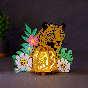 May include: A 3D papercraft lamp featuring a leopard design. The lamp is illuminated and has a jungle theme with green leaves and flowers. The lamp is made of paper and is a decorative item.