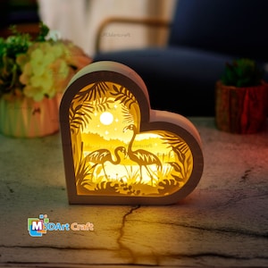 May include: A white wooden heart-shaped night light with a cutout design of two flamingos standing in a tropical scene with palm trees and a full moon. The night light is illuminated from behind, casting a warm glow.