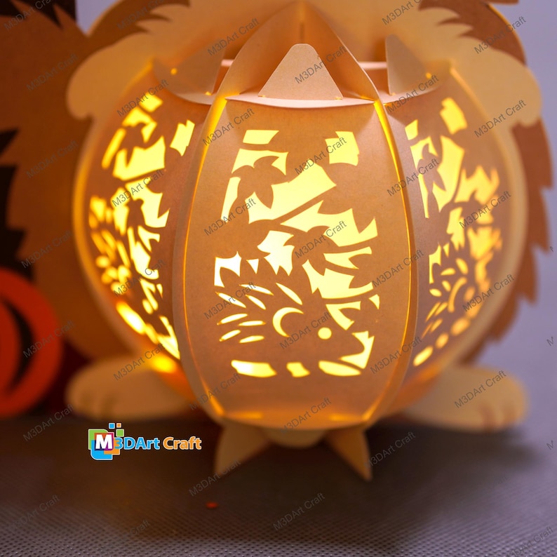 May include: A paper lantern in the shape of a hedgehog, with intricate cut-out designs. The lantern is illuminated from within, casting a warm, inviting glow. The design includes leaves and a hedgehog silhouette. The product is from M3DArt Craft.
