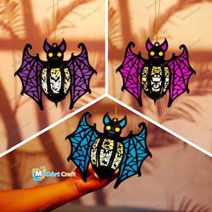 May include: Three hanging bat ornaments with intricate cut-out designs. The bats are purple, pink, and blue with a white glow in the center. The bats are hanging from a string.