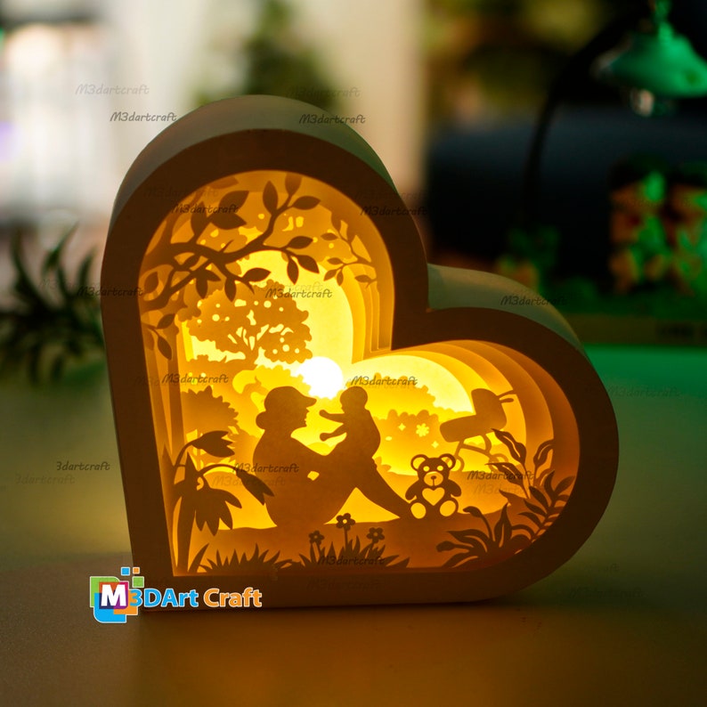 May include: A white heart-shaped wooden lamp with a 3D layered design. The lamp features a mother and child sitting in a garden with a teddy bear and a swing set. The lamp is illuminated with warm white light.