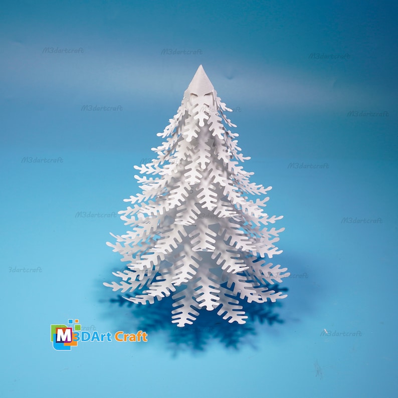 Peut inclure: Un arbre de No&euml;l en papier blanc avec un design 3D. L'arbre est compos&eacute; de nombreuses couches de papier d&eacute;coup&eacute;es et pli&eacute;es pour cr&eacute;er un aspect r&eacute;aliste. L'arbre est pos&eacute; sur une surface bleue.