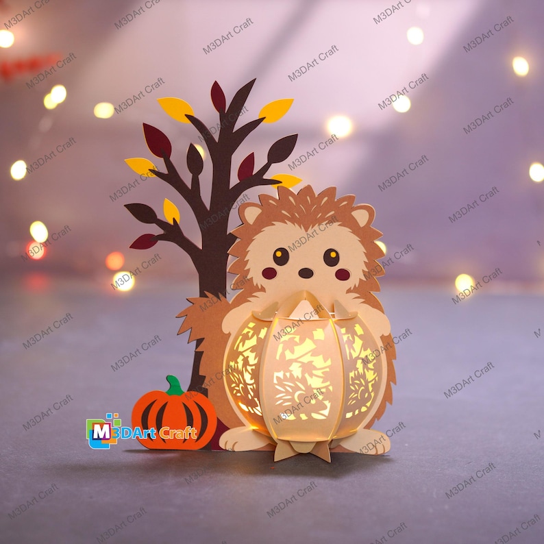 May include: A papercraft hedgehog holding a lantern, with a tree and pumpkin. The lantern has a floral design and emits a warm glow. The tree has brown branches and yellow and maroon leaves. The pumpkin is orange.