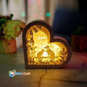 May include: A white heart-shaped wooden night light with a cut-out design of a cat and dog sitting on a bench. The design also includes flowers, leaves, and the word "Love" in the center. The light is illuminated from behind, creating a warm glow.
