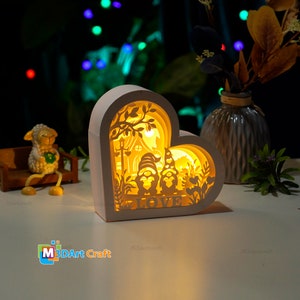 Puede incluir: Lámpara de madera blanca en forma de corazón con un diseño de corte 3D de dos gnomos de pie frente a una casa con un árbol y la palabra "LOVE" en la parte inferior. La lámpara está encendida y proyecta un brillo cálido.