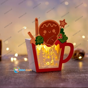 K&ouml;nnte beinhalten: Eine beleuchtete Weihnachts-Holztasse mit einem Lebkuchenmann-Design. Die Tasse ist rot und wei&szlig; mit einem ausgeschnittenen Design einer Winterszene mit B&auml;umen, einem Haus und einem Reh.