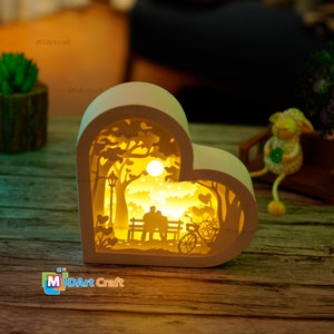 May include: A white heart-shaped wooden lamp with a cutout design of a couple sitting on a bench in a park with a bicycle and trees. The lamp is lit up with warm yellow light.