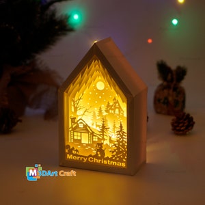 May include: A white papercraft house with a light inside, depicting a winter scene with a cabin, trees, a snowman, and deer. The text "Merry Christmas" is visible at the bottom of the house.