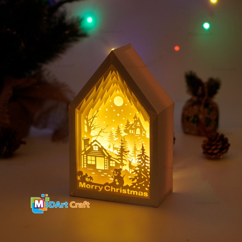 May include: A white papercraft house with a light inside, depicting a winter scene with a cabin, trees, a snowman, and deer. The text "Merry Christmas" is visible at the bottom of the house.