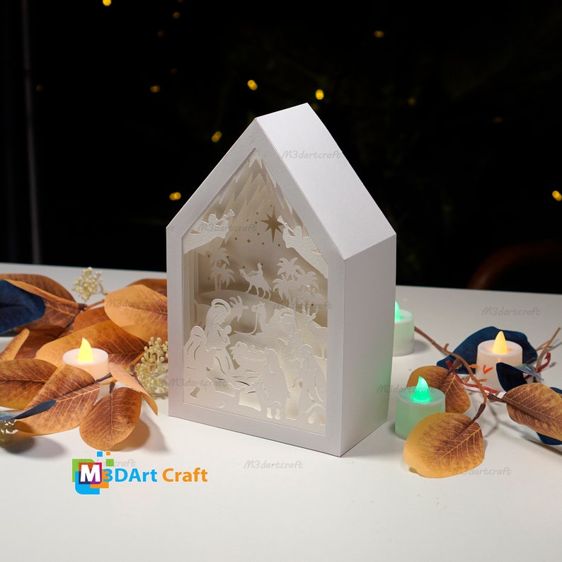 May include: White papercraft nativity scene with a cutout design of the baby Jesus, Mary, Joseph, and animals. The scene is illuminated by a light source, creating a warm glow. The papercraft is in the shape of a house and has the text "M3DArt Craft" on the bottom.