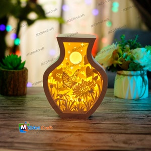 May include: A white ceramic vase with a cut-out design of a butterfly, flowers, and a moon. The vase is illuminated from within, creating a warm glow.