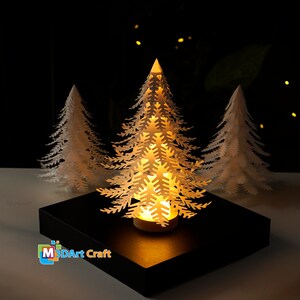 Peut inclure: Trois sapins de No&euml;l en papier blanc avec des d&eacute;coupes complexes, le sapin du milieu est &eacute;clair&eacute; par une lumi&egrave;re blanche chaude, le tout sur une base carr&eacute;e noire. La base porte l'inscription "M3DArt Craft".