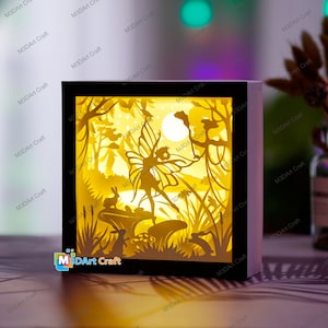 May include: A black wooden frame with a yellow and white paper cut-out design of a fairy with wings, a moon, and rabbits in a forest setting. The frame has a light inside that illuminates the design.