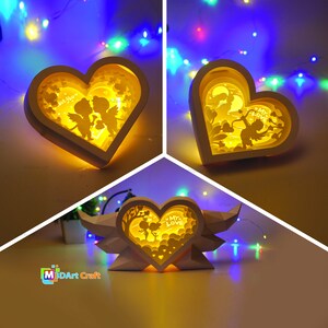 May include: Three white papercraft heart-shaped lamps with intricate cut-out designs. The lamps are illuminated from within, casting a warm glow. The designs feature a couple of angels, a heart, and the text "My Love".