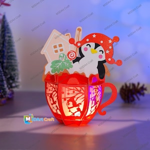 Puede incluir: Una linterna de papel roja y blanca con forma de taza con un ping&uuml;ino que lleva un gorro rojo y sostiene un bast&oacute;n de caramelo en su interior. La taza est&aacute; decorada con una casa de jengibre y hojas verdes. La linterna est&aacute; iluminada desde el interior, creando un brillo c&aacute;lido.
