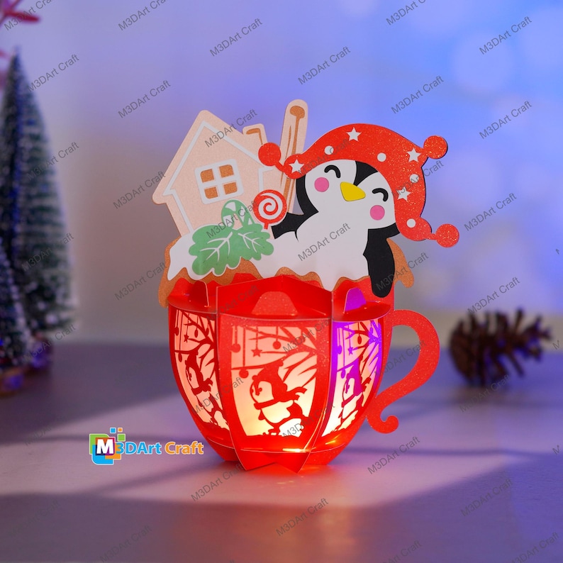 Puede incluir: Una linterna de papel roja y blanca con forma de taza con un ping&uuml;ino que lleva un gorro rojo y sostiene un bast&oacute;n de caramelo en su interior. La taza est&aacute; decorada con una casa de jengibre y hojas verdes. La linterna est&aacute; iluminada desde el interior, creando un brillo c&aacute;lido.