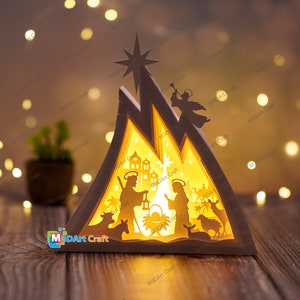 May include: A white wooden nativity scene with a star on top. The scene is illuminated from behind, creating a warm glow. The figures of Mary, Joseph, and the baby Jesus are visible, along with an angel and animals.