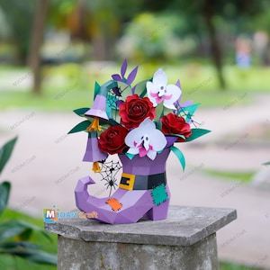 Puede incluir: Un arreglo de papel con un jarrón en forma de bota de bruja morada lleno de rosas rojas, flores blancas con forma de fantasma y flores moradas con forma de campana. La bota está decorada con un cinturón negro y parches naranjas y verdes.