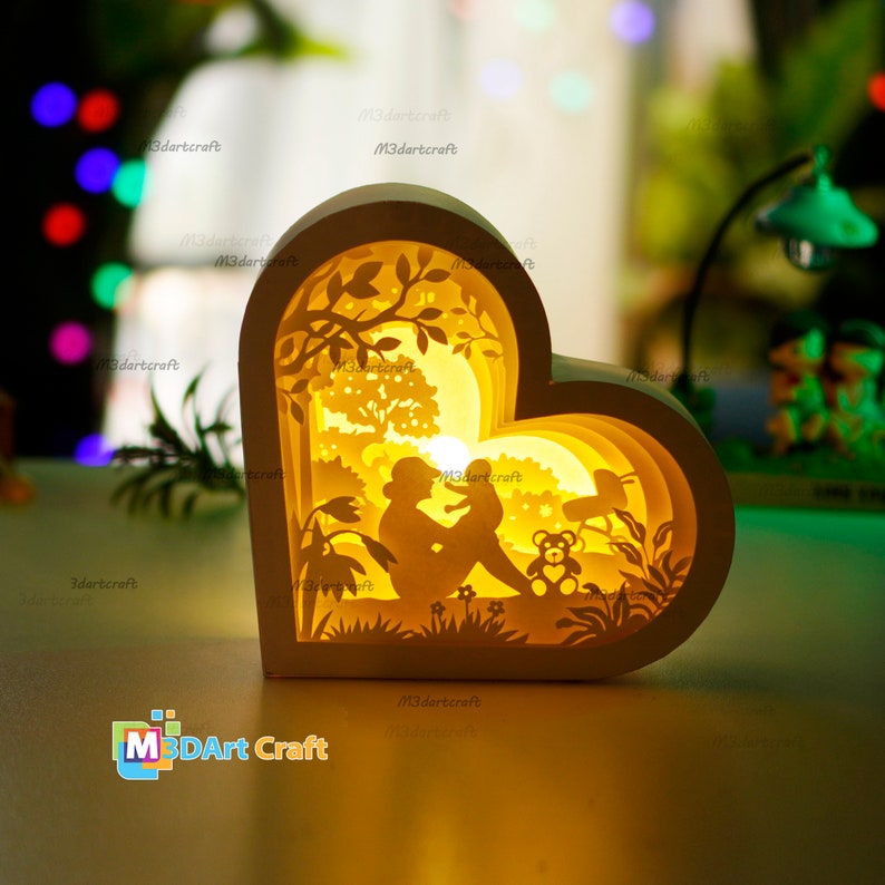 May include: A white wooden heart-shaped lamp with a cutout design of a mother and child sitting in a forest. The lamp is illuminated from within, casting a warm glow.