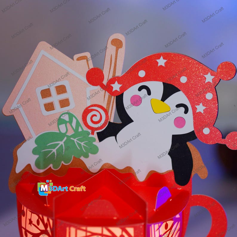 Puede incluir: Un ping&uuml;ino rojo y blanco con un gorro de Pap&aacute; Noel sostiene un bast&oacute;n de caramelo y asoma por un mug rojo con una casa de jengibre en la parte superior. El mug est&aacute; decorado con glaseado blanco y purpurina.